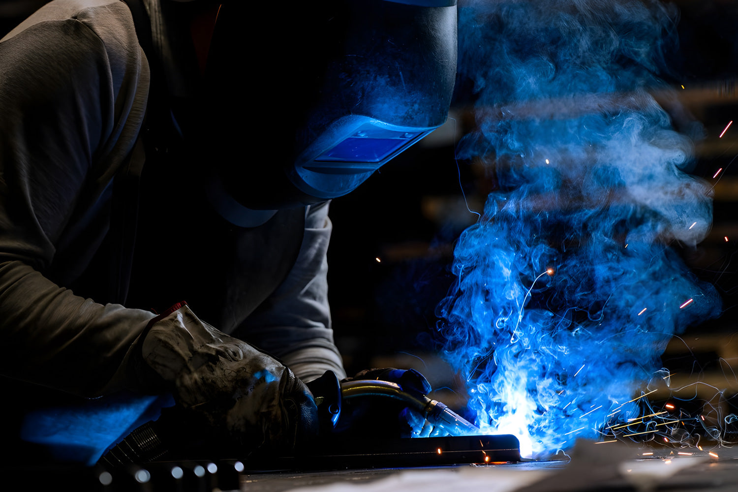 A welder wearing protective gear works on a metal piece, with bright blue sparks and smoke rising from the welding process. The scene is dimly lit, highlighting the intense blue light and providing a dramatic contrast.