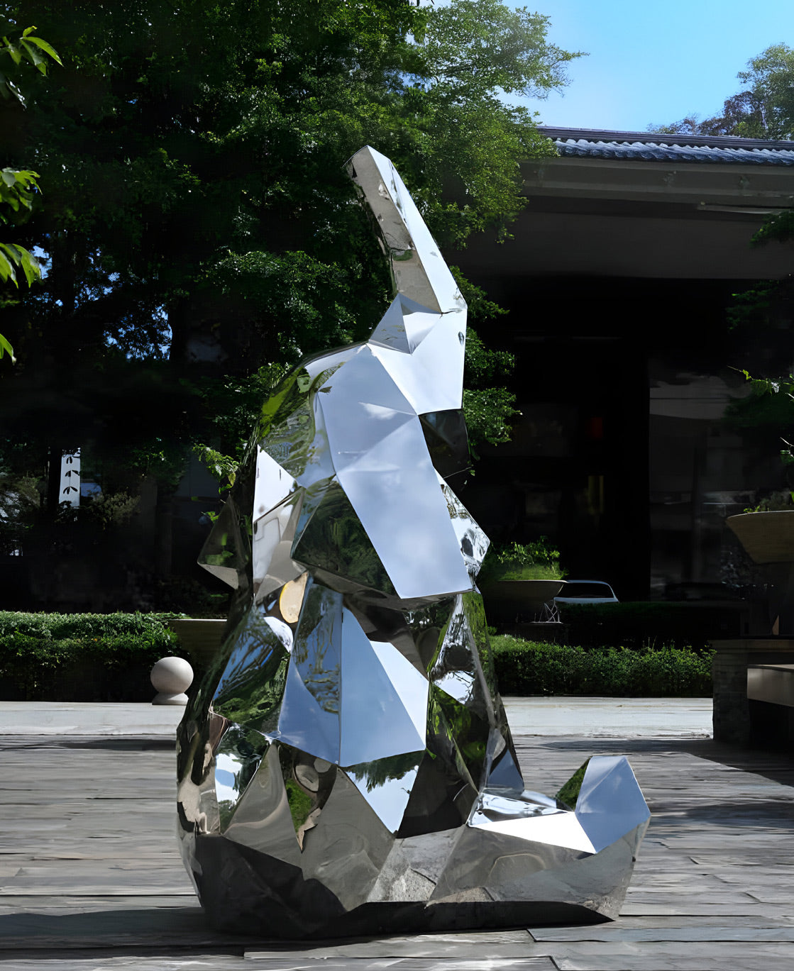 A large, reflective geometric sculpture shaped like an elephant is situated outdoors. The background features greenery and a building. The sculptures mirrored surfaces reflect its surroundings.