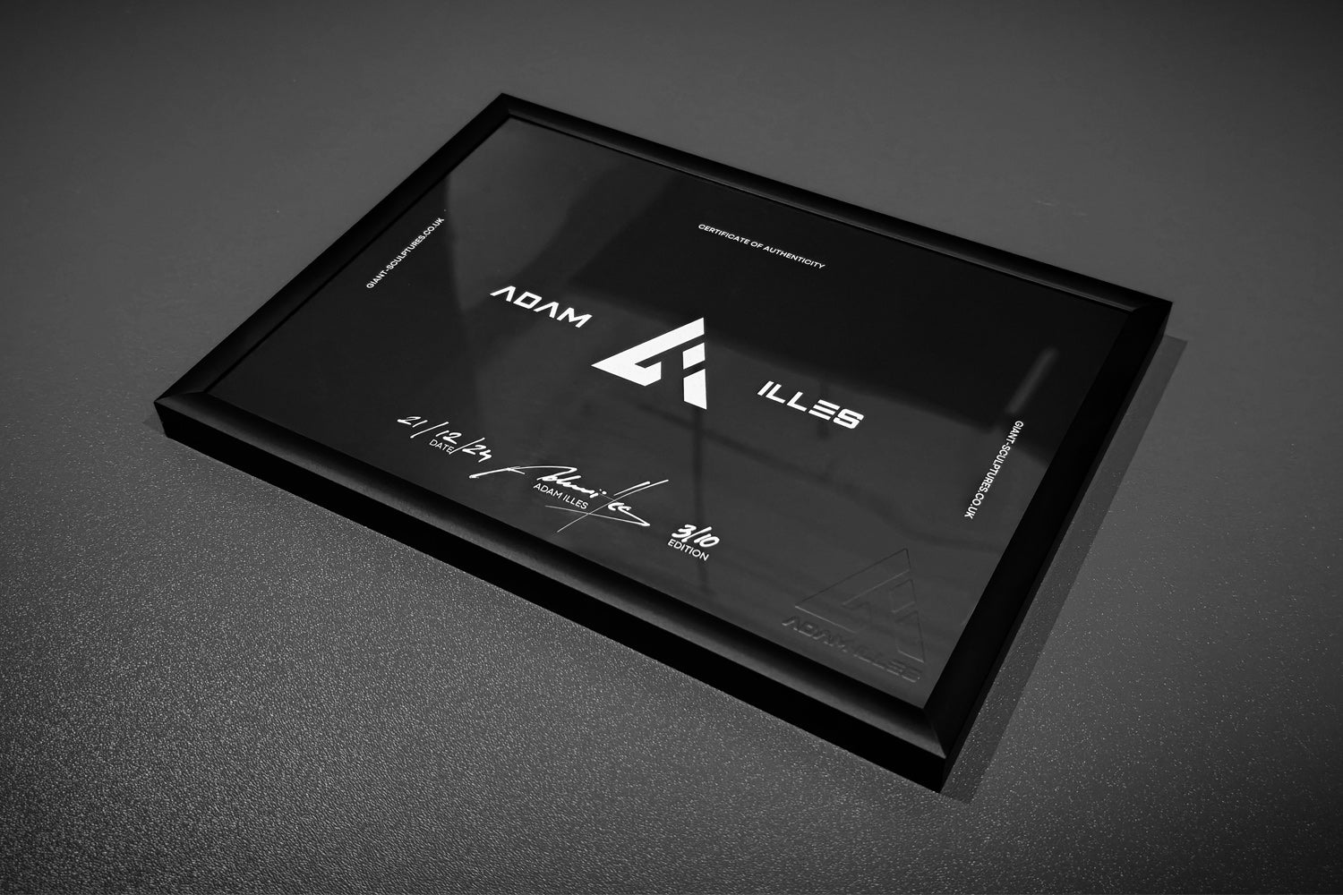 A black plaque with white text is displayed. It features the name Adam Illes and a logo resembling the letter A. Several signatures and the date 21 July 2023 are visible. The plaque is framed in black and placed on a dark surface.