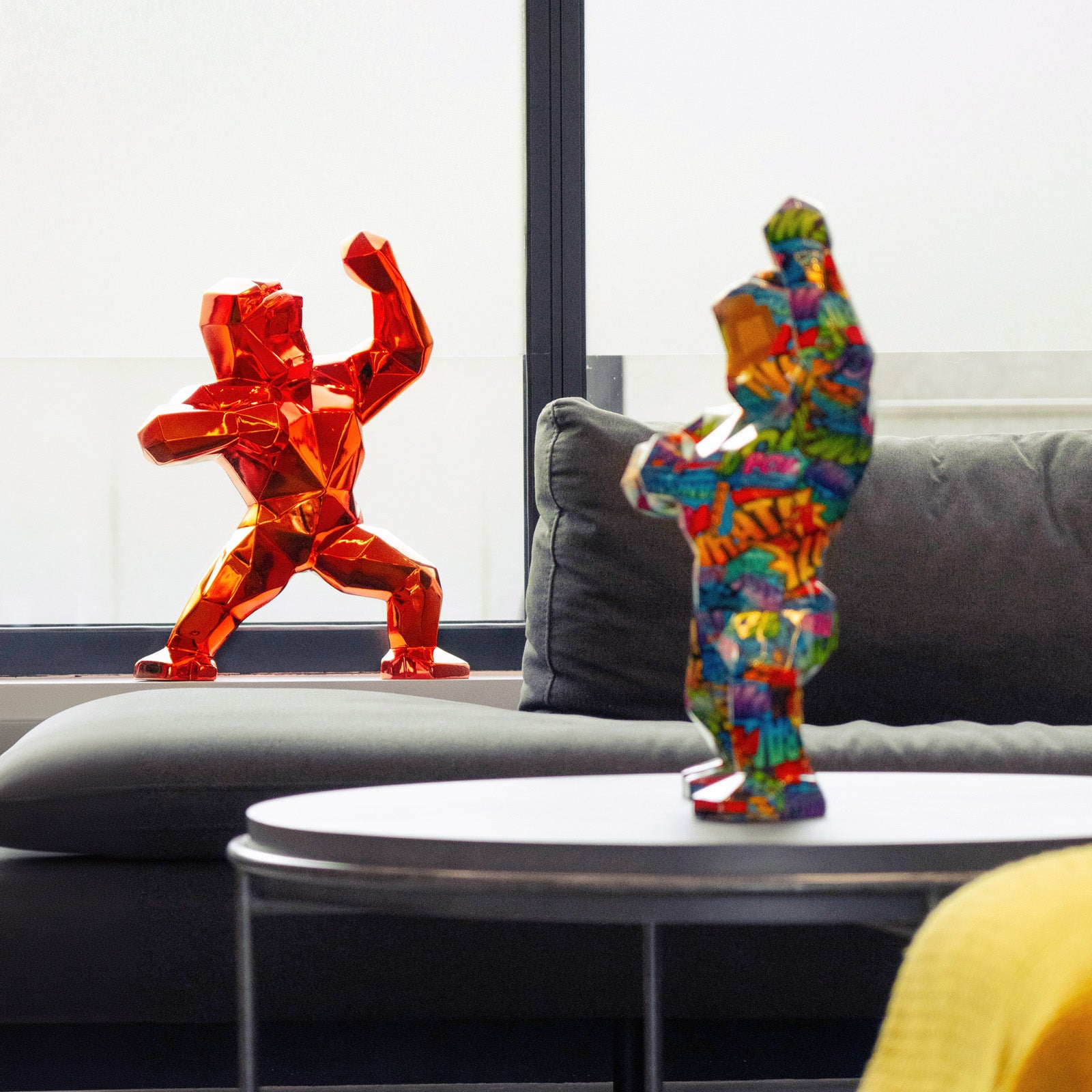 Two colorful gorilla sculptures, one in red and the other with a comic-style pattern, stand on a table and sofa. They face each other in a bright room with large windows, creating a modern and artistic atmosphere.