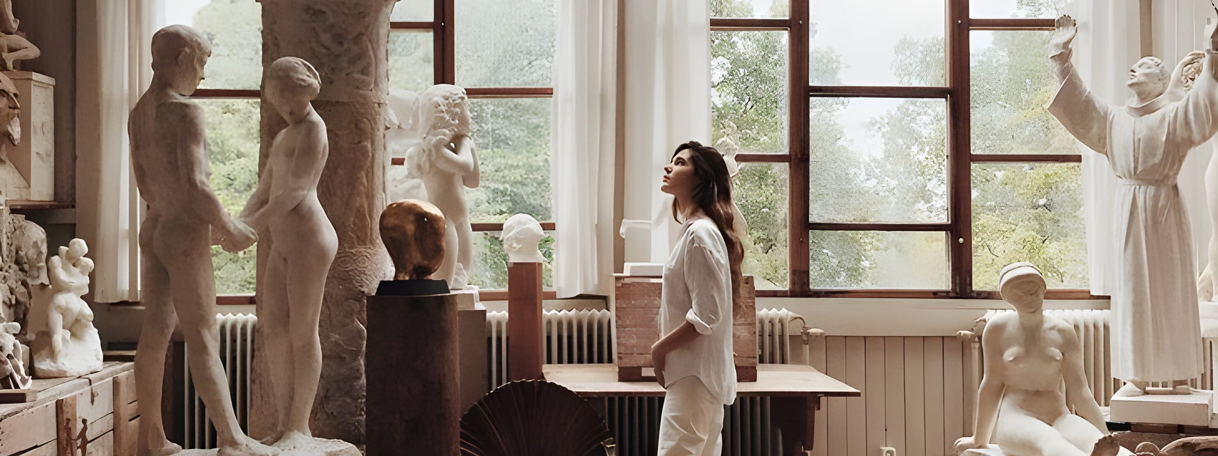 A woman stands in an art studio surrounded by various sculptures. She looks up at a statue, with large windows letting in natural light. The studio has a serene and creative atmosphere, with greenery visible outside.