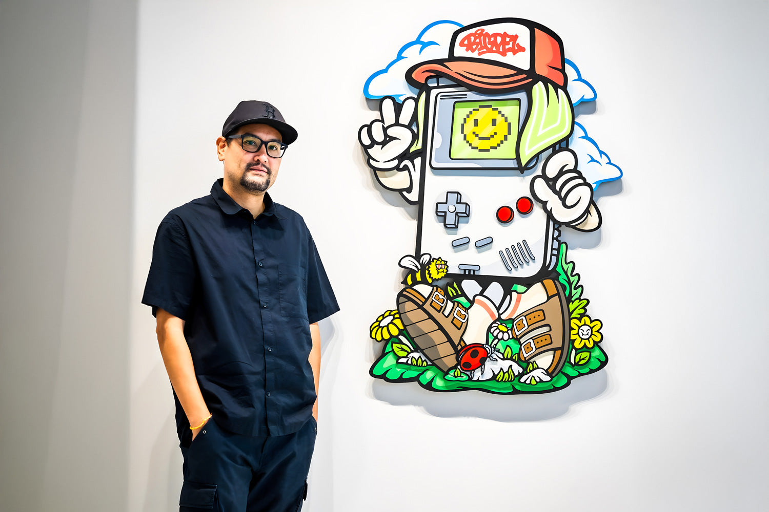 A person in black clothing and glasses stands next to a colorful wall art piece depicting a cartoon Game Boy wearing sneakers, a cap, and a smiley face sticker. The background features clouds and flowers.