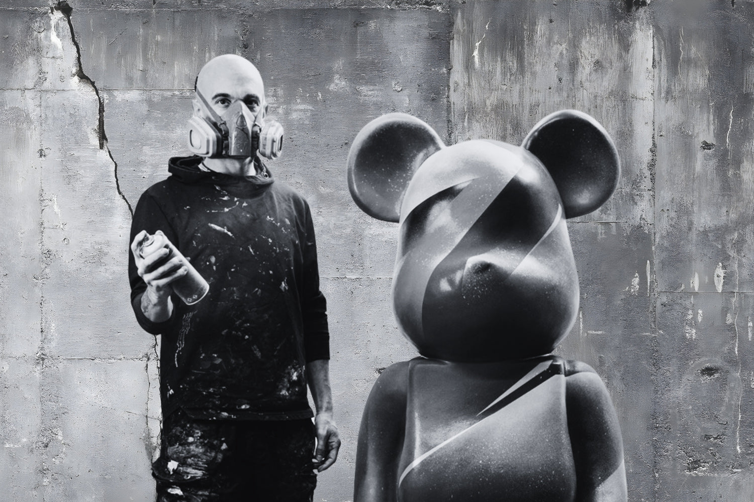 A man in a gas mask holds a spray can, standing next to a large abstract sculpture with a bear-like head. The background features a textured concrete wall with a visible crack. The scene is in black and white.