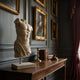 The Draped Male Marble Torso Sculpture (120cm) by Giant Sculptures stands on a wooden table with books, candlesticks, and framed portraits against a dark-paneled wall in a room with rich drapes and classical decor.