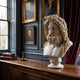 The Zeus Marble Bust Sculpture - 55cm by Giant Sculptures sits on a wooden cabinet in a classic study with blue paneled walls, framed portraits, red drapes, and stacks of old books.