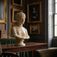 The Classical Female Marble Bust Sculpture - 70cm by Giant Sculptures is showcased on a wooden table in an elegant room with framed portraits, books, and large windows letting in natural light.