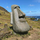 The Moai Easter Island Head Stone Sculpture - 100cm by Giant Sculptures stands on grassy terrain with hills, palm trees, and the ocean in the background beneath a blue sky, evoking Easter Island.