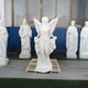 A collection of white statues is indoors. In the center, the Skyward Gaze Angel Marble Outdoor Sculpture by Giant Sculptures showcases wings spread wide, encircled by smaller figures, set against a blue and glass-paneled wall, evoking an outdoor garden centerpiece.