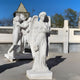 In the foreground stands the Giant Sculptures Tender Heart Angel Marble Outdoor Sculpture - 200cm, showcasing a white angel with flowers and wings folded back. The serene outdoor scene is complemented by nearby statues of three figures holding hands under a clear blue sky.