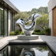 The Wisp Silver Abstract Steel Sculpture - 200cm by Giant Sculptures stands on a circular pedestal in a modern courtyard, its sleek form reflected in a still rectangular pool with trees and greenery behind.