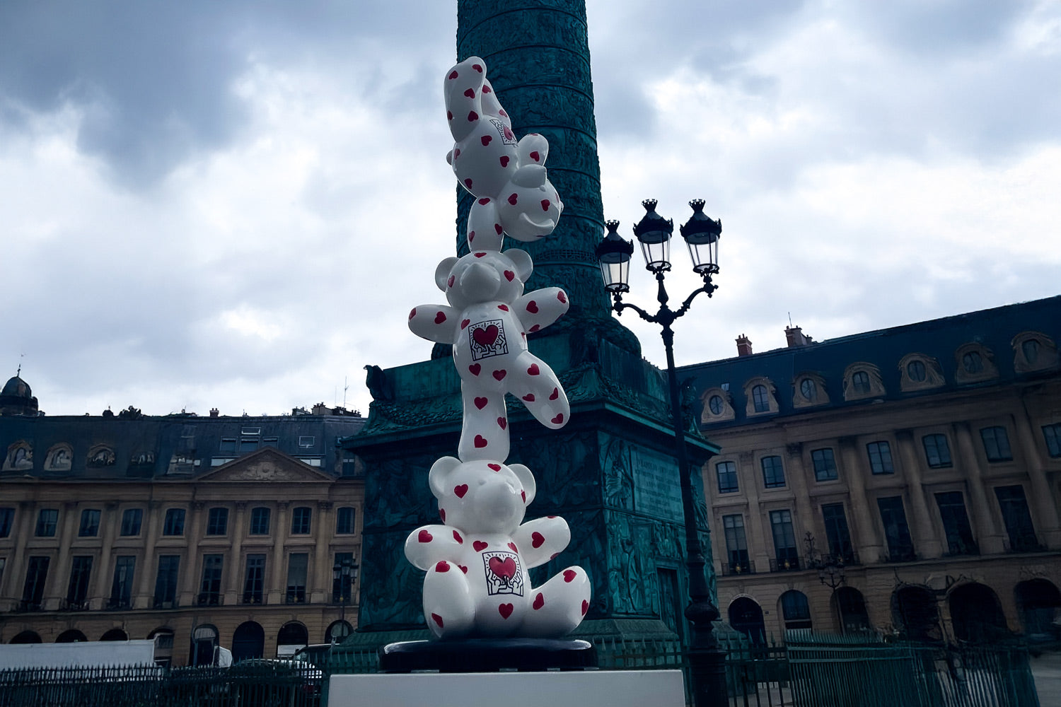 A stack of white teddy bears with red hearts is positioned in front of a tall, ornate green column. The sky is overcast, and historic buildings are visible in the background, alongside an ornate streetlamp.