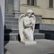 The Kneeling Prayer Angel Marble Outdoor Sculpture - 120cm by Giant Sculptures, with its intricate wings and peaceful expression, adorns a stone platform before the building steps, embodying tranquility in the daylight.