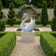 The Radiant Swan White Marble Outdoor Sculpture (100cm) by Giant Sculptures stands on a stone pedestal in a formal garden, framed by manicured hedges, tall trees, gravel paths, and an arched brick wall with climbing roses.