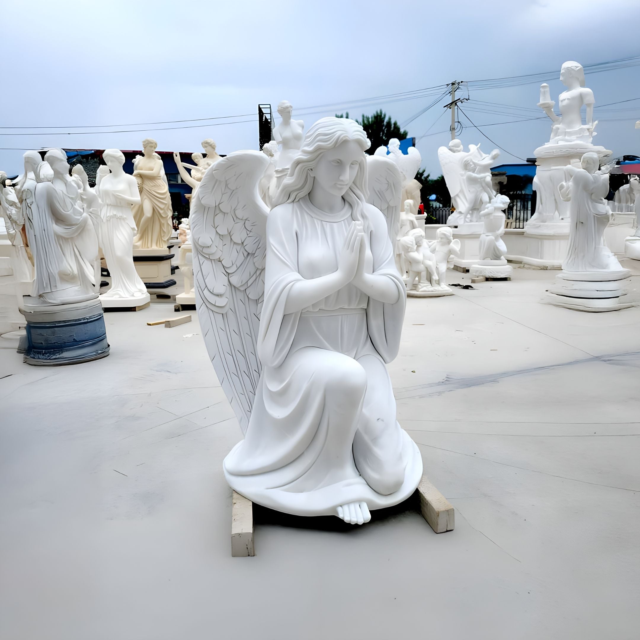 The Kneeling Serenity Angel Marble Outdoor Sculpture from Giant Sculptures, standing at 150cm, depicts a kneeling angel with clasped hands in prayer. Complete with detailed wings and a serene expression, it is set among other marble statues on a smooth, light-toned surface.