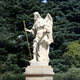 The 200cm Armoured Guardian Angel Marble Outdoor Sculpture by Giant Sculptures stands amidst tall evergreen trees, featuring an angel in motion with wings, a long staff, flowing robes, and protective armor on a stone pedestal.