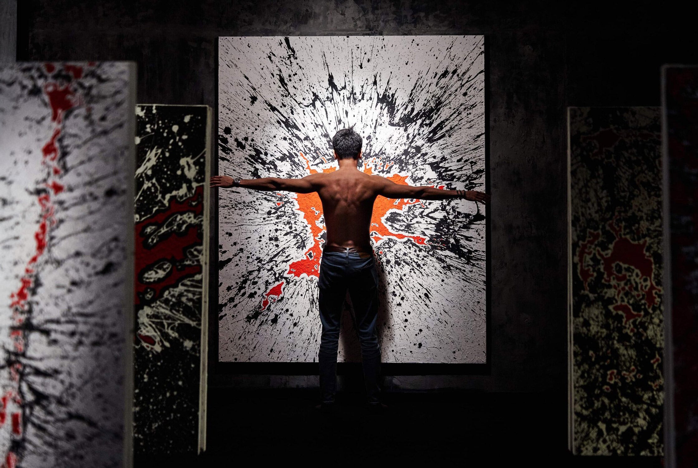 A shirtless person stands with arms outstretched in front of a large, abstract painting featuring splatters of black, white, and red. Several similar artworks are partially visible around them in a dimly lit space.