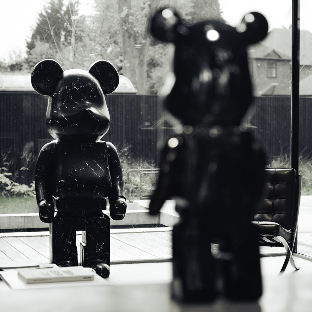 Two large, black bear-shaped sculptures stand indoors near a window with a view of a yard. One sculpture is in the foreground with its back to the viewer, and the other faces forward, creating a mirrored effect. Trees and a chair are visible outside.