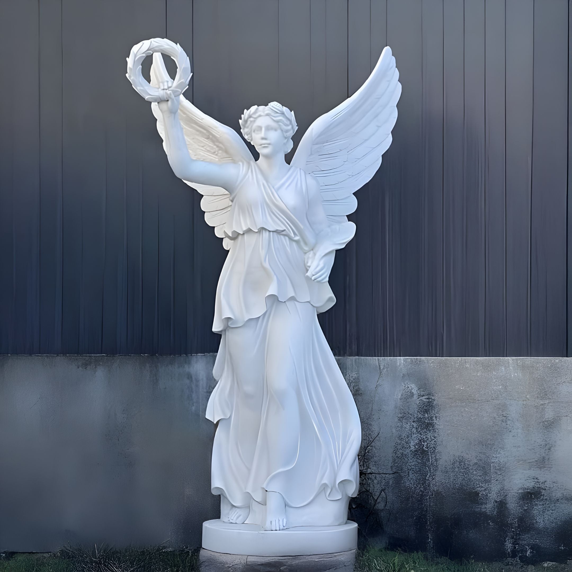 The Victory Wreath Angel Marble Outdoor Sculpture by Giant Sculptures, standing 220cm, features a winged angel in flowing robes gracefully holding a victory wreath. Set against a concrete and dark paneled background, this white marble piece is the ideal garden statue exuding timeless beauty.