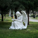 The Reading Grace Angel Marble Outdoor Sculpture by Giant Sculptures shows a serene angel reading a book on grass. This 120cm contemplative figure is set against tall trees and a blurred road, enhancing the garden memorials tranquil ambiance.