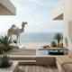 A modern terrace with the Majestic Camel White Marble Outdoor Sculpture (160cm) by Giant Sculptures, desert plants, a small pool, and beige seating forms a tranquil minimalist oasis under a clear sky.
