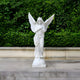 The Crowned Serenity Angel Marble Outdoor Sculpture by Giant Sculptures, standing 200cm tall, depicts a graceful angel with wings and crossed arms on a stone platform in front of a lush green hedge.