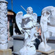 Amidst a clear blue sky, the Giant Sculptures Guardian Staff Angel Marble Outdoor Sculpture stands at 190cm; one winged angel plays a trumpet while the other looks contemplative, shielding its eyes.