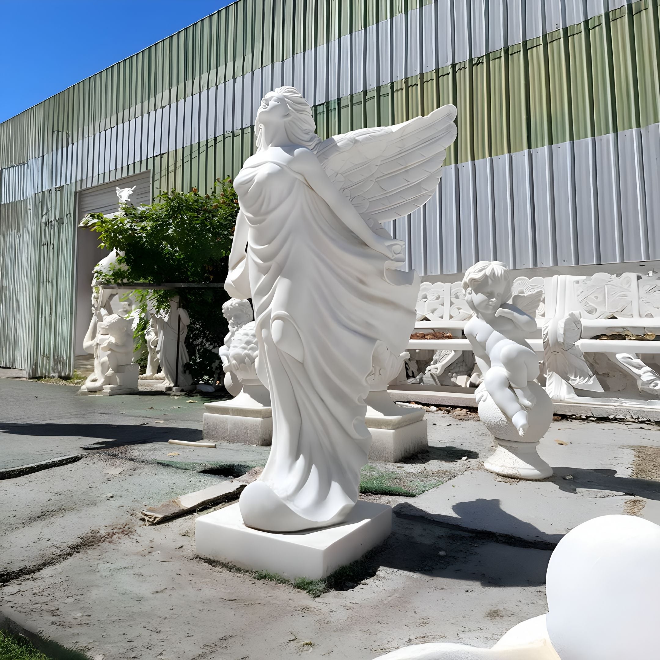 The Billowing Gown Angel Marble Outdoor Sculpture by Giant Sculptures, measuring 200cm, stands gracefully against a corrugated metal backdrop. Accompanied by a cherub-like figure, it adds an ethereal touch to the garden amidst lush greenery.