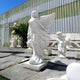 The Billowing Gown Angel Marble Outdoor Sculpture by Giant Sculptures, measuring 200cm, stands gracefully against a corrugated metal backdrop. Accompanied by a cherub-like figure, it adds an ethereal touch to the garden amidst lush greenery.