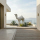 The Elegant Camel Marble Outdoor Sculpture by Giant Sculptures (160cm) is set among agave plants on a patio with beige walls, offering views of the ocean and distant mountains beneath a hazy sky.