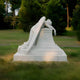 The Weeping Angel Marble Outdoor Sculpture by Giant Sculptures, standing 150cm tall, features a white marble angel with wings draped over a pedestal, gazing down in mourning. This poignant garden memorial is set amidst lush greenery and framed by tall trees.