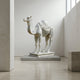 The Desert Voyager Camel Marble Outdoor Sculpture (160cm) by Giant Sculptures stands elegantly on a pedestal in a minimalist, light-filled gallery with neutral walls and floor—a striking piece of desert camel decor.