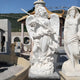 The garden features a 190cm Defender Staff Angel Marble Outdoor Sculpture by Giant Sculptures, depicting a serene white marble angel with wings, flowing robes, detailed hair, and a flute. A second sculpture is partially visible on the right amidst a gazebo and rolling green hills.