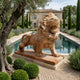 The Giant Lion II Yellow Marble Outdoor Sculpture - 240cm by Giant Sculptures stands beside a tree and trimmed bushes, overlooking a reflecting pool in an elegant garden with a grand estate—a striking luxury outdoor décor piece.