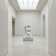 The Kneeling Serenity Angel Marble Outdoor Sculpture (150cm) by Giant Sculptures, in white marble with clasped hands, stands at the center of a minimal, light-filled gallery with pale walls and a skylight above.