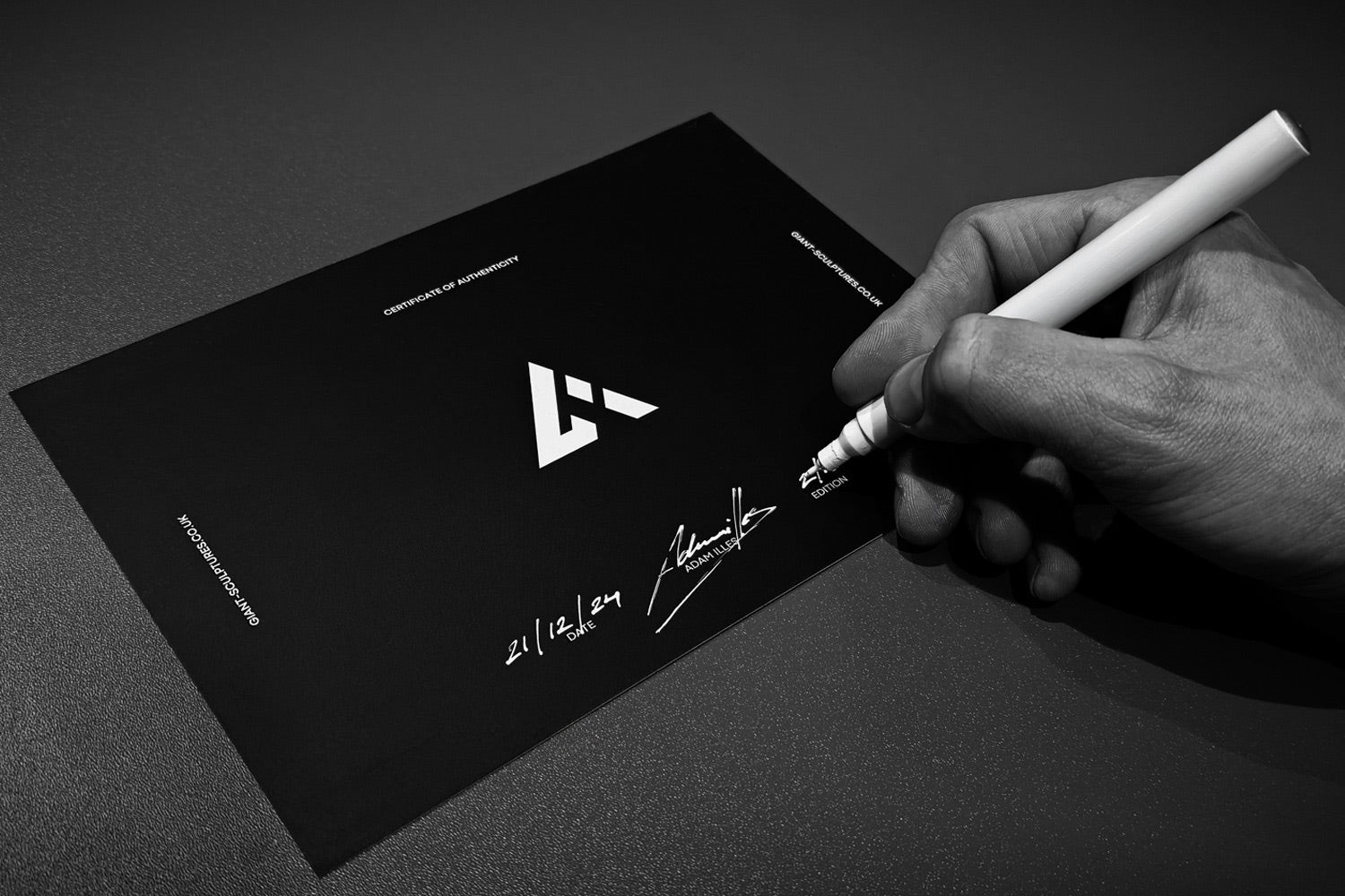 A hand holds a white pen, signing a black document titled Certificate of Ownership with a geometric logo. The signature is in cursive. The date reads 21 | 12 | 24 with additional text beneath. The image is in black and white.