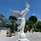 The Dove Release Peace Angel Marble Outdoor Sculpture by Giant Sculptures stands 280cm tall, depicting a white angel with wings holding a dove, surrounded by trees and statues under the sun, alongside a forklift near a building.