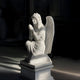 The 180cm Crowned Prayer Angel Marble Outdoor Sculpture by Giant Sculptures features a white marble angel kneeling on a pedestal with clasped hands, long hair, and intricately carved wings. Soft lighting enhances its ethereal presence against dark surroundings.