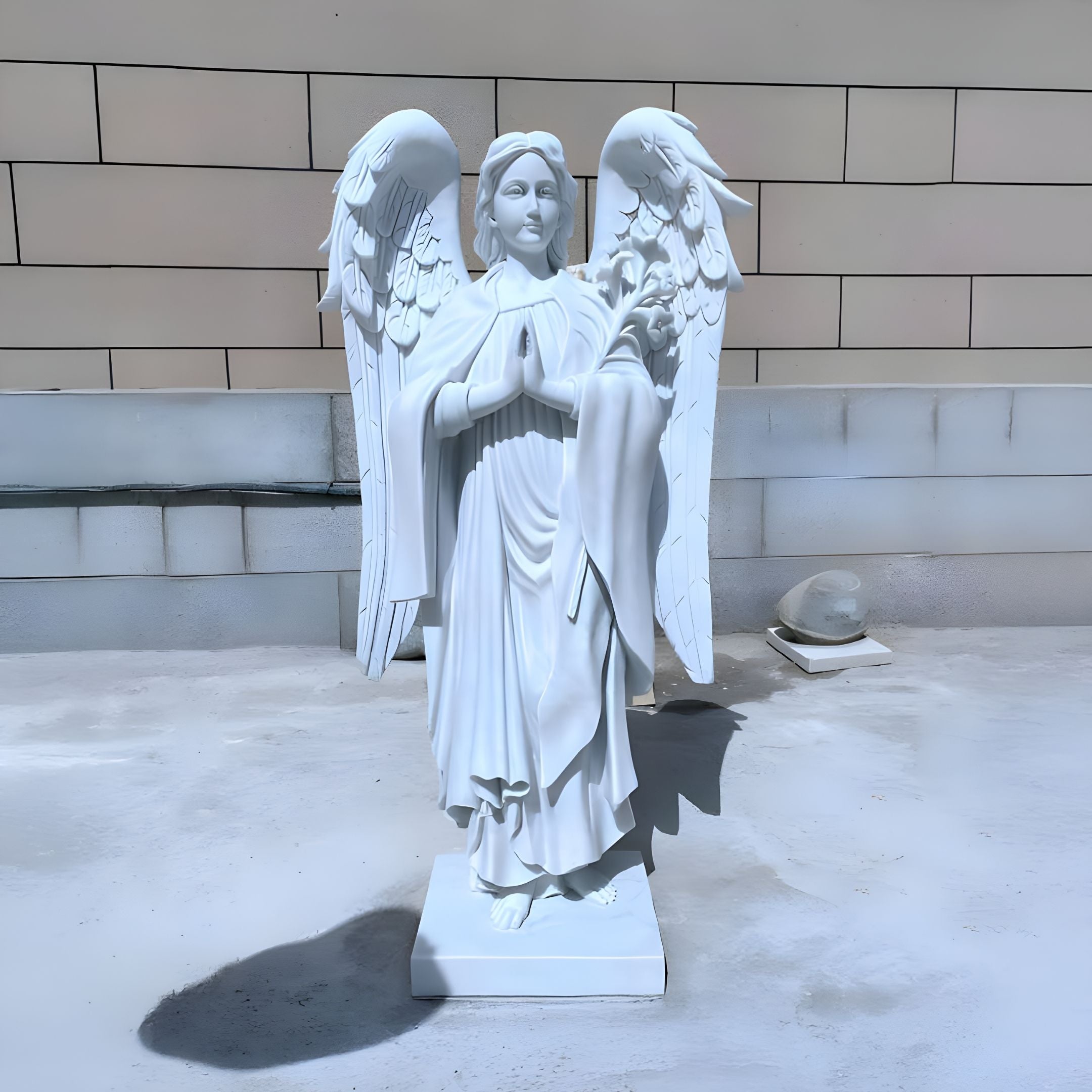 The Prayerful Laurel Angel Marble Outdoor Sculpture - 130cm by Giant Sculptures features a white marble angel with large wings and prayerful hands. Set on a light stone surface against a tiled wall, this serene garden statue casts an elegant shadow.