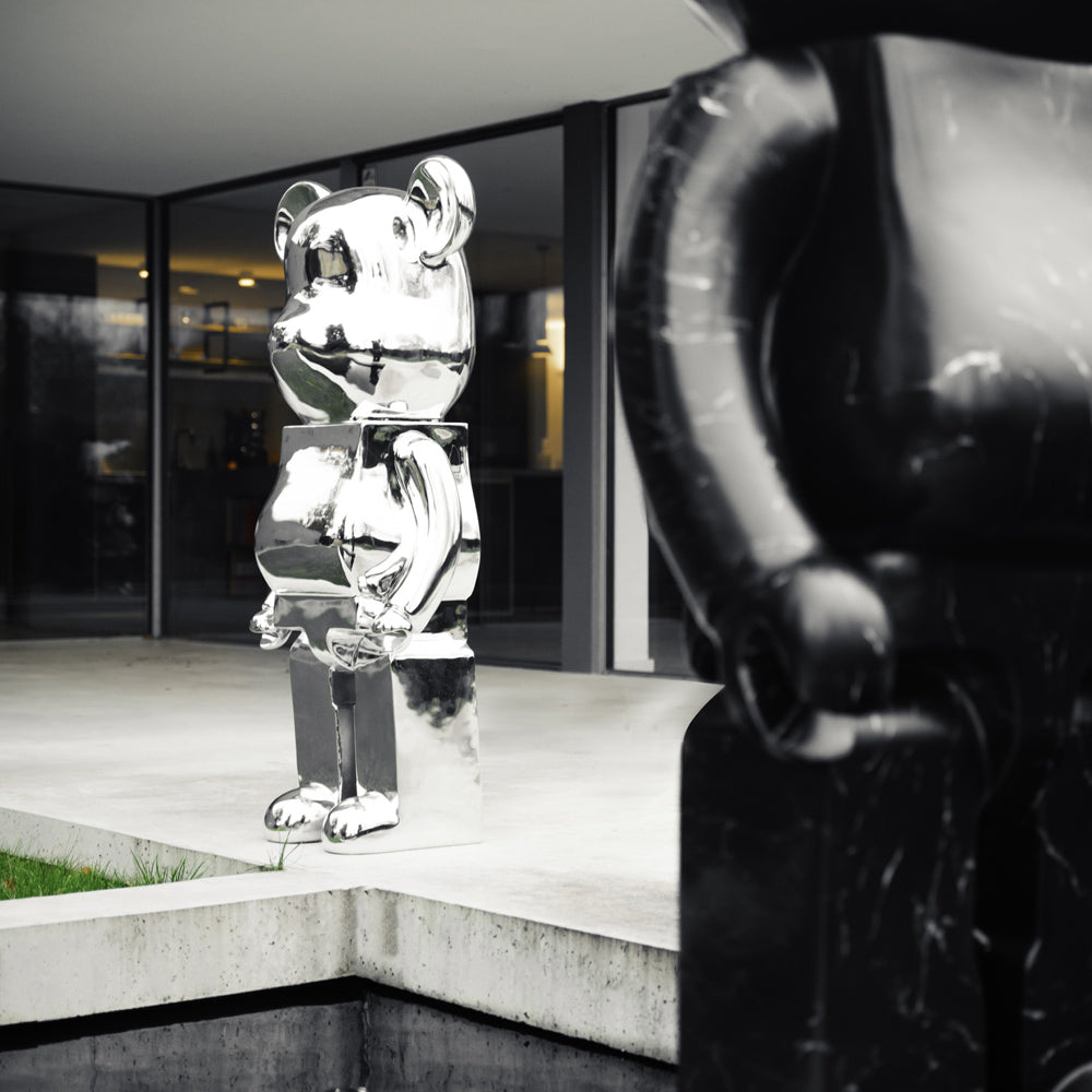 A shiny metallic bear sculpture stands on the corner of a modern buildings patio, mirrored by another similar black figure in the foreground. The area features minimalistic architecture with large glass windows.