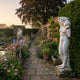 Standing Draped Woman Marble Sculpture 160cm by Giant Sculptures, posed with an urn on her shoulder beside a yew hedge in a walled rose garden at dusk.