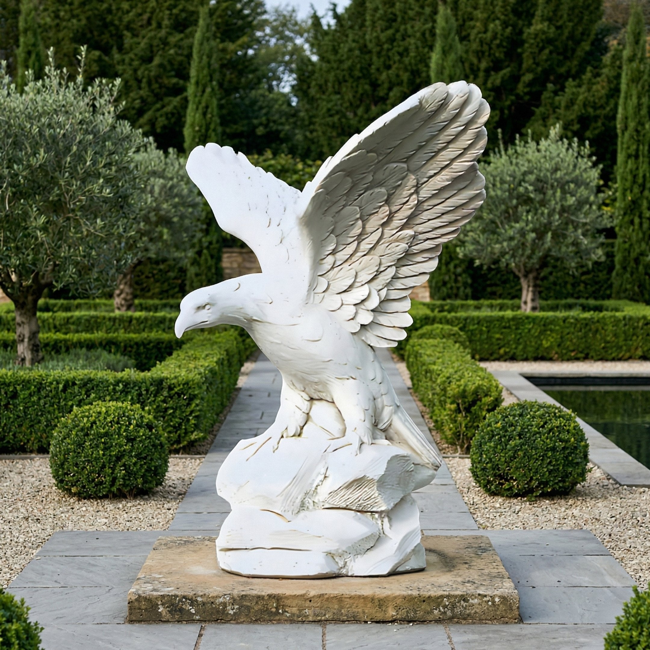 The Giant Sculptures Rock-Perched Eagle Marble Outdoor Sculpture (120cm) stands on a pedestal in a formal garden with trimmed bushes, trees, gravel paths, and a reflecting pool nearby.