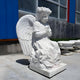 The 100cm Blossom Crown Cherub Marble Outdoor Sculpture by Giant Sculptures features a kneeling cherub with wings and a floral crown. This serene statue is placed outdoors near a building with blue and red paneling, surrounded by planters.