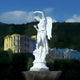 The Dancing Harvest Angel Marble Outdoor Sculpture by Giant Sculptures elegantly poses in the foreground of green hills and distant buildings beneath a clear blue sky.