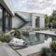 A modern concrete house with large glass windows overlooks a reflecting pool, featuring the 65cm Walking Polar Bear White Marble Outdoor Sculpture by Giant Sculptures. Lush landscaping surrounds the serene, minimalist courtyard.