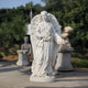 The Rose-Gathering Angel Marble Outdoor Sculpture by Giant Sculptures, 180cm tall, features detailed hand-carved wings and flowing robes as it holds flowers in a garden setting enhanced by blurred green foliage and other outdoor sculptures.