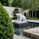 The Seated Polar Bear White Marble Outdoor Sculpture - 50cm by Giant Sculptures stands on a pedestal in a reflecting pool, surrounded by manicured greenery, trimmed bushes, and a modern stone garden.