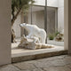The Polar Bear Family White Marble Outdoor Sculptures (180cm) by Giant Sculptures stand on a rock indoors near large windows, with an olive tree and minimalist landscaping visible outside.