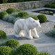 The Arctic Polar Bear White Marble Outdoor Sculpture - 50cm by Giant Sculptures stands on a paved area in a landscaped garden with green bushes, ornamental grasses, gravel, and a pond with lily pads.