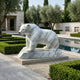 The Polar Bear White Marble Outdoor Sculpture - 50cm by Giant Sculptures stands on a stone base beside a pool in a formal garden, surrounded by trimmed hedges, gravel, olive trees, and modern architecture.