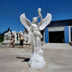 The Torch-Bearing Angel Marble Outdoor Sculpture - 220cm by Giant Sculptures stands gracefully before a blue and white industrial building, with other sculptures in the background under a clear blue sky.